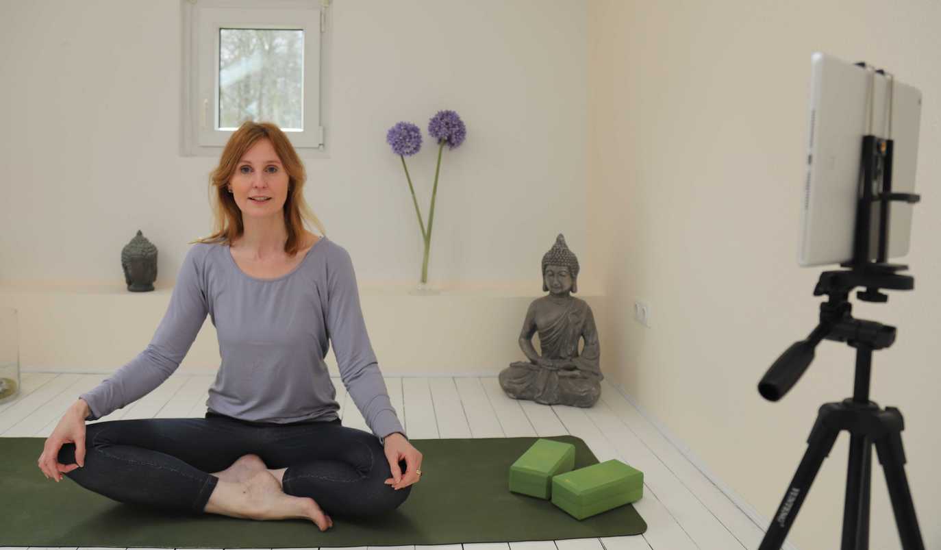 Eine Frau sitzt im Schneidersitz auf einer Yogamatte, vor ihr steht ein Stativ mit einem Tablet.