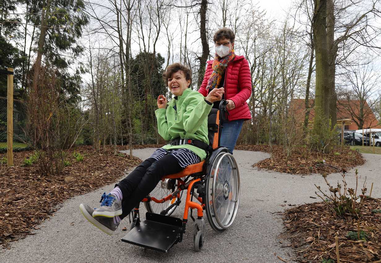 Eine freudig lachende Frau in einem Rollstuhl und wird von einer anderen Frau über einen Weg geschoben.